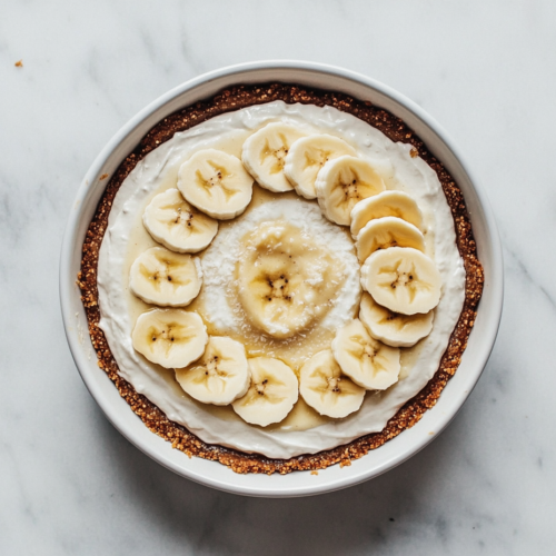 This image shows a vegan banana cream pie presented in a round white baking dish. The pie features a creamy filling made with whipped coconut cream, vanilla, and powdered sugar, topped with perfectly arranged banana slices.