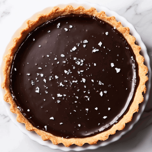 This image shows a rich and glossy vegan dark chocolate tart in a round dish, with the smooth chocolate filling perfectly set in a golden crust, ready to serve as an indulgent dessert.