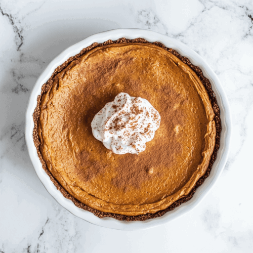 This image shows a vegan pumpkin pie in a round dish with a rich, spiced filling in the center. A dollop of creamy vegan coconut whipped cream sits elegantly on top, ready to be enjoyed.