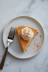 This image shows a perfectly sliced piece of vegan pumpkin pie topped with a generous dollop of vegan coconut whipped cream, highlighting the creamy filling and flaky crust.