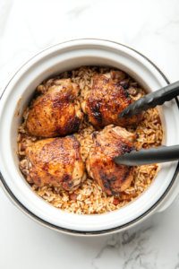 This image shows golden-browned chicken thighs being placed on top of the seasoned rice and vegetable mixture in the slow cooker, ready to cook slowly to perfection.