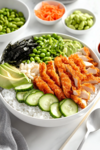 This image shows a bowl being carefully assembled with rice, sliced chicken katsu, fresh cucumber, avocado, and pickled ginger for a vibrant and delicious meal.