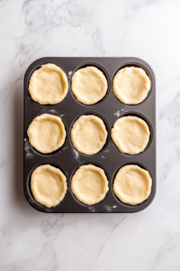 This image shows a top-down view of a muffin tin with pieces of refrigerated biscuit dough pressed into each cup, creating a base for the mini chicken pot pies before filling.