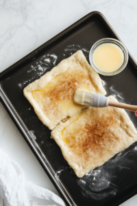 This image shows a pastry brush spreading melted butter on crescent dough, followed by a generous sprinkling of cinnamon sugar to add sweetness and flavor.