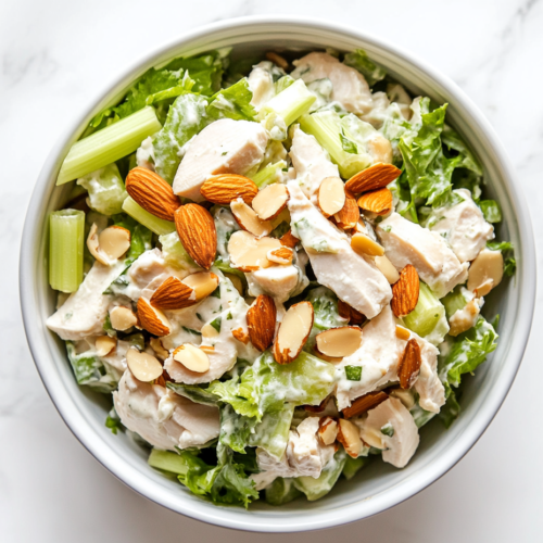 This image shows a white bowl filled with creamy Chicken Salad, made with cooked shredded or chopped chicken, toasted almonds, and chopped celery, creating a fresh and crunchy texture.