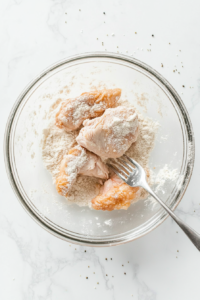 This image shows a piece of buttermilk-marinated chicken being dredged in a bowl of seasoned flour, forming the crispy coating for frying.