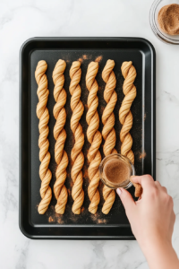 This image shows twisted crescent dough strips lined up on a baking sheet, being coated with an extra layer of cinnamon sugar for a crispy, flavorful finish.