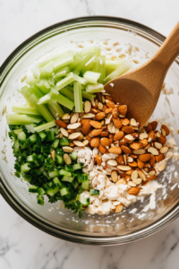 This image shows shredded chicken, toasted almonds, and chopped celery being added to a mixing bowl, ready to be coated with the dressing.