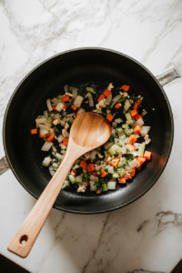 This image shows diced onions, celery, minced garlic, and sliced carrots sizzling in a pot, being sautéed to release their natural sweetness and aroma for a flavorful chicken vegetable stew.