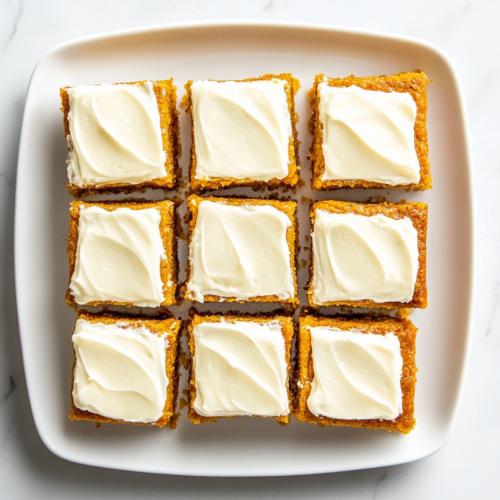 This image shows a batch of homemade pumpkin bars, cut into perfect squares and topped with a thick layer of smooth cream cheese frosting, making for a delicious autumn treat.