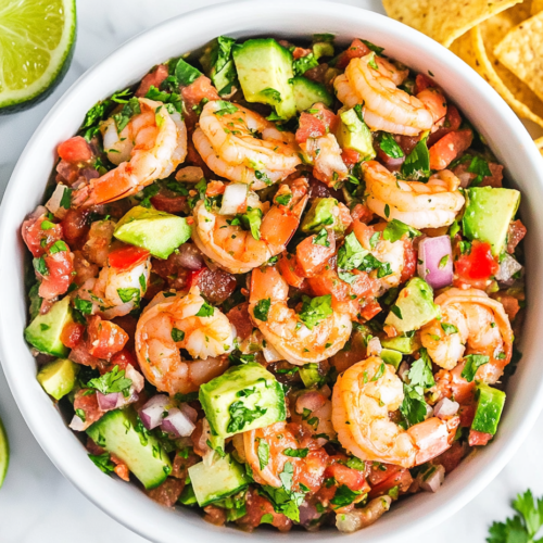 This image shows a white bowl filled with freshly made Avocado Shrimp Salsa, featuring diced cucumbers, tomatoes, onions, and avocados, mixed with seasoned shrimp and garnished with cilantro for a bright and refreshing dish.