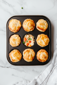 This image shows a top-down view of a muffin tin on the counter, filled with freshly baked mini chicken pot pies, their golden-brown crusts crisp and slightly puffed as they cool.