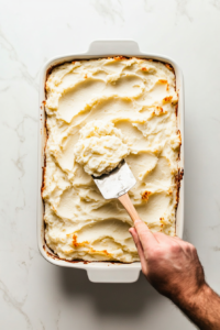 This image shows a thick layer of creamy mashed potatoes being spread evenly over the cooked turkey meatloaf in the baking dish.
