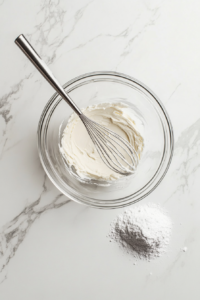 This image shows a mixing bowl with softened cream cheese, butter, powdered sugar, and vanilla being whipped together to create a smooth and creamy frosting.