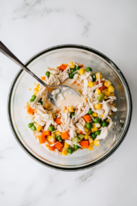 This image shows a top-down view of a mixing bowl filled with ingredients for mini chicken pot pie filling, including cream of chicken soup, shredded chicken, and vegetables, being combined with a spoon.