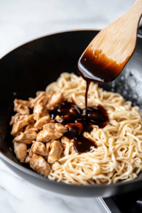 This image shows the cooked chicken, noodles, and stir-fry sauce being added to the wok, coming together for a delicious lo mein dish