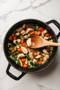 This image shows a large spoon stirring together chicken, vegetables, and broth in a simmering pot, ensuring that all the flavors meld together perfectly for a hearty stew.