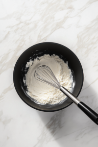 This image shows a wooden spoon stirring flour and oil together in a pot, forming the base of a roux, which will add depth and richness to the gumbo.