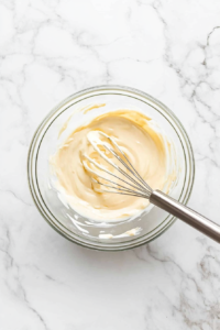 This image shows a rich, glossy sauce being whisked together in a bowl, combining honey, mayonnaise, and condensed milk for the perfect balance of sweetness and creaminess.