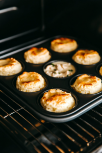 This image shows a top-down view of a muffin tin inside an oven, where the mini chicken pot pies are baking to golden perfection with their biscuit dough crusts rising around the creamy filling.