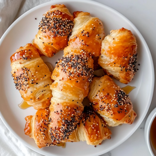 This image shows a plate of golden brown, crispy pigs in a blanket topped with sesame seeds, served with a small bowl of maple syrup on the side.