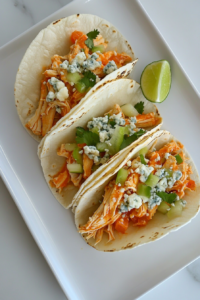 This image shows a plate of Buffalo chicken tacos, garnished with blue cheese, scallions, and a side of lemon wedges, ready to be served with extra sauce for dipping.