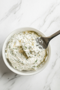 This image shows a rich and creamy veggie dip being transferred from a mixing bowl into a white serving bowl, ready to be served.