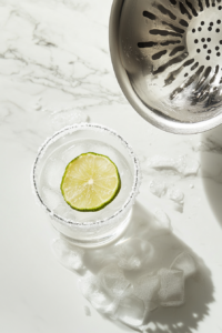 This image shows a freshly shaken vodka margarita being poured into a salt-rimmed glass filled with ice, then garnished with a lemon slice for a bright, citrusy finish.
