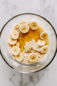 This image shows melted butter, vanilla extract, and an egg being added to the mashed bananas, forming the wet mixture for the cookies.