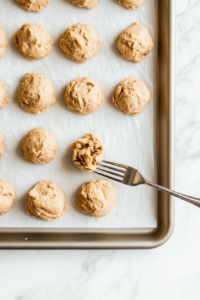 This image shows a spoon gently flattening cookie dough balls on a baking sheet, ensuring they bake evenly with a slightly crisp edge.