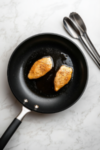 This image shows a coated chicken breast sizzling in a pan, cooking to a golden brown crust on one side.
