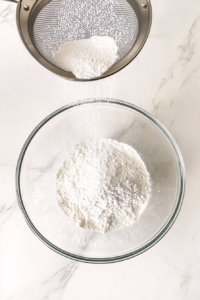 This image shows flour, baking soda, and other dry ingredients being sifted together into a mixing bowl, ensuring an even and smooth cookie dough texture.
