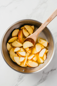 Apple slices gently cooking in a pot with fruit juice, releasing their natural sweetness and softening for a rich, homemade pie filling.