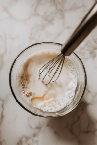 This image shows warm milk being gradually poured into a whisked egg mixture, tempering the eggs to prevent curdling.