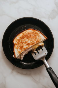This image shows a spatula flipping a turkey melt sandwich on a skillet, ensuring even browning and melting the cheese perfectly inside.