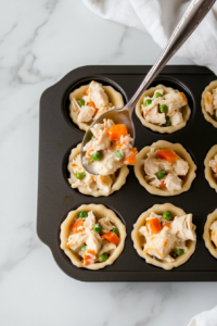 This image shows a top-down view of a spoon scooping the creamy chicken pot pie filling into the biscuit dough cups inside a muffin tin, preparing them for baking.