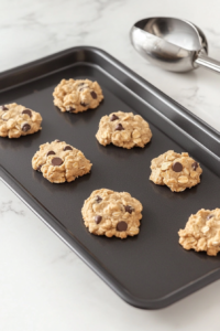 This image shows spoonfuls of cookie dough being dropped onto a greased baking sheet, ready to be baked into golden, chewy Cowboy Cookies.