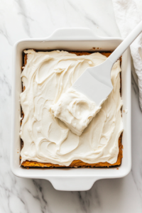 This image shows a spatula spreading a thick, velvety layer of cream cheese frosting over the cooled pumpkin bars, creating a luscious and even coating.