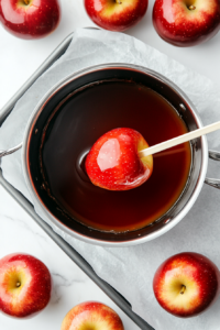 This image shows whole apples being dipped into the hot candy syrup, coating them evenly in a smooth, shiny layer of red candy.