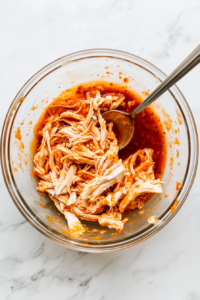This image shows shredded chicken being tossed in a bowl of spicy Buffalo sauce, ensuring every piece is coated with bold, tangy flavor.