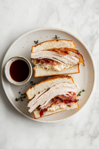This image shows a turkey melt sandwich served on a plate, with crispy bacon, melted cheese, and fresh vegetables, ready to be enjoyed.
