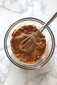 This image shows dry ingredients, including flour, baking soda, salt, and cinnamon, being whisked together in a bowl for an evenly blended mixture.