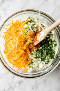This image shows a bowl of cream cheese being mixed with shredded cheese, herbs, and seasonings to create a flavorful cheese ball mixture.