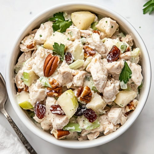 This image shows a delicious bowl of chicken salad made with canned chicken, creamy salad dressing, pickle relish, diced apples, chopped pecans, sliced celery, and raisins, creating a flavorful and textured dish.
