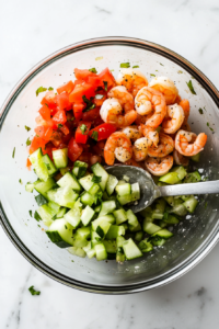 This image shows a large mixing bowl filled with shrimp, tomatoes, and other fresh vegetables being combined for a vibrant and refreshing dish.