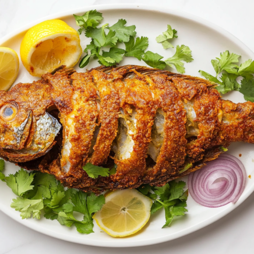 This image shows a whole fish fried to crispy, golden perfection, garnished with fresh chopped cilantro, and served on a round white plate with onion rings and a half-cut lemon on the side.