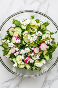 This image shows the process of crumbling goat cheese over the cucumber radish salad, adding a creamy and tangy touch to the dish.