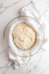 This image shows the dough resting in a bowl, covered, to allow it to hydrate properly before shaping into Gulab Jamuns.