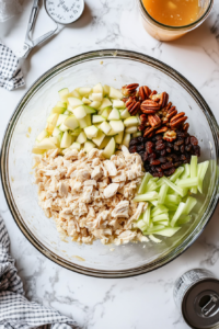 This image shows a large mixing bowl filled with canned chicken, creamy dressing, pickle relish, diced apples, chopped pecans, sliced celery, and raisins, as all the ingredients are combined to make a flavorful chicken salad.