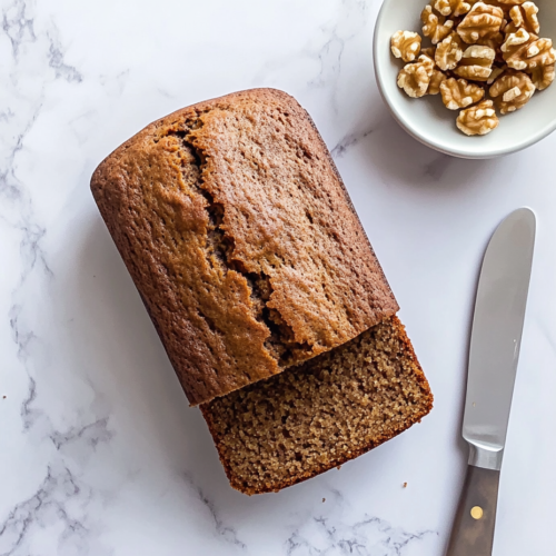 This image shows a freshly baked brown banana bread loaf with a moist interior, placed on a white marble slab. One slice is cut with a knife resting on the side, and a small white bowl filled with walnuts sits nearby.